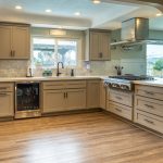 L-shaped kitchen with greige shaker cabinets, wine fridge, window over the sink, and a range with stainless hood