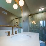 Brushed gold faucet on a white vanity top under globe lights, with a glass shower enclosure in the background
