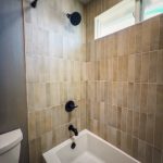 Tub and shower combo with vertical stacked tile, matte black shower fixtures, and a frosted window