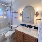 Wood vanity with white countertop, navy tile backsplash, arched mirror, and wall sconces beside floating wood shelves