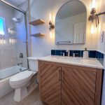 Remodeled La Mesa bathroom with chevron wood vanity, arched mirror, blue tile backsplash, floating shelves, and glass tub doors