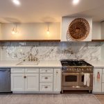 White shaker kitchen with full-height marble-look slab backsplash, stainless range, and floating wood shelf