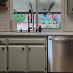 Matte black pull-down faucet over a white sink, framed by subway tile, a garden-view window, and sage green cabinets
