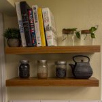 Natural wood floating shelves styled with cookbooks, glass storage jars, plants, and a cast iron teapot
